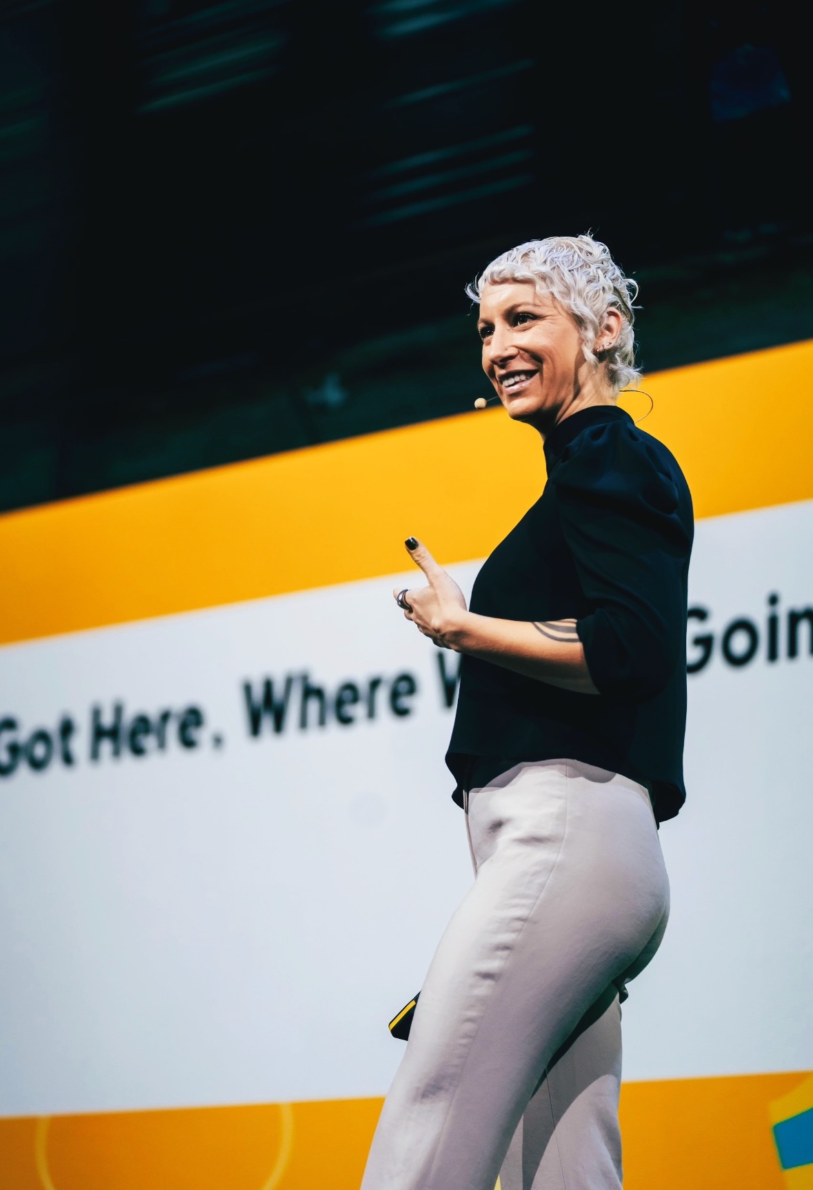 Lily Ray on stage smiling and looking back at the audience, wearing a black top and white pants, with a bright orange presentation slide visible behind her