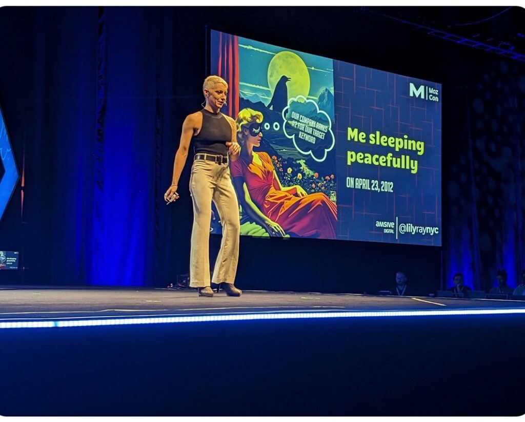 Lily Ray speaking on the MozCon stage with a large illuminated X logo backdrop and audience members visible in the foreground